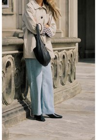 Personne portant une veste beige, un jean large bleu clair et des chaussures noires pointues, tenant un sac tissé noir, debout près d'une rambarde en pierre ornée.