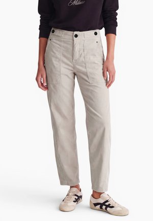 Personne portant un pantalon beige clair à jambe droite avec des poches plaquées à l’avant et des détails de boutons noirs, assorti à des baskets beige et noires.