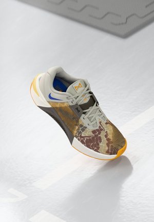 But sportowy z kamuflażowym wzorem, żółto-biała podeszwa, szare sznurowadła, niebieskie logo Nike w kształcie łyżwy oraz logo na języku, spoczywający na jasnej powierzchni.