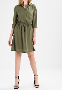 Oliwkowa sukienka koszulowa z zapinanym przodem, rękawami o długości 3/4, elastycznym pasem i gładką, zwiewną fakturą. Czarne szpilki.