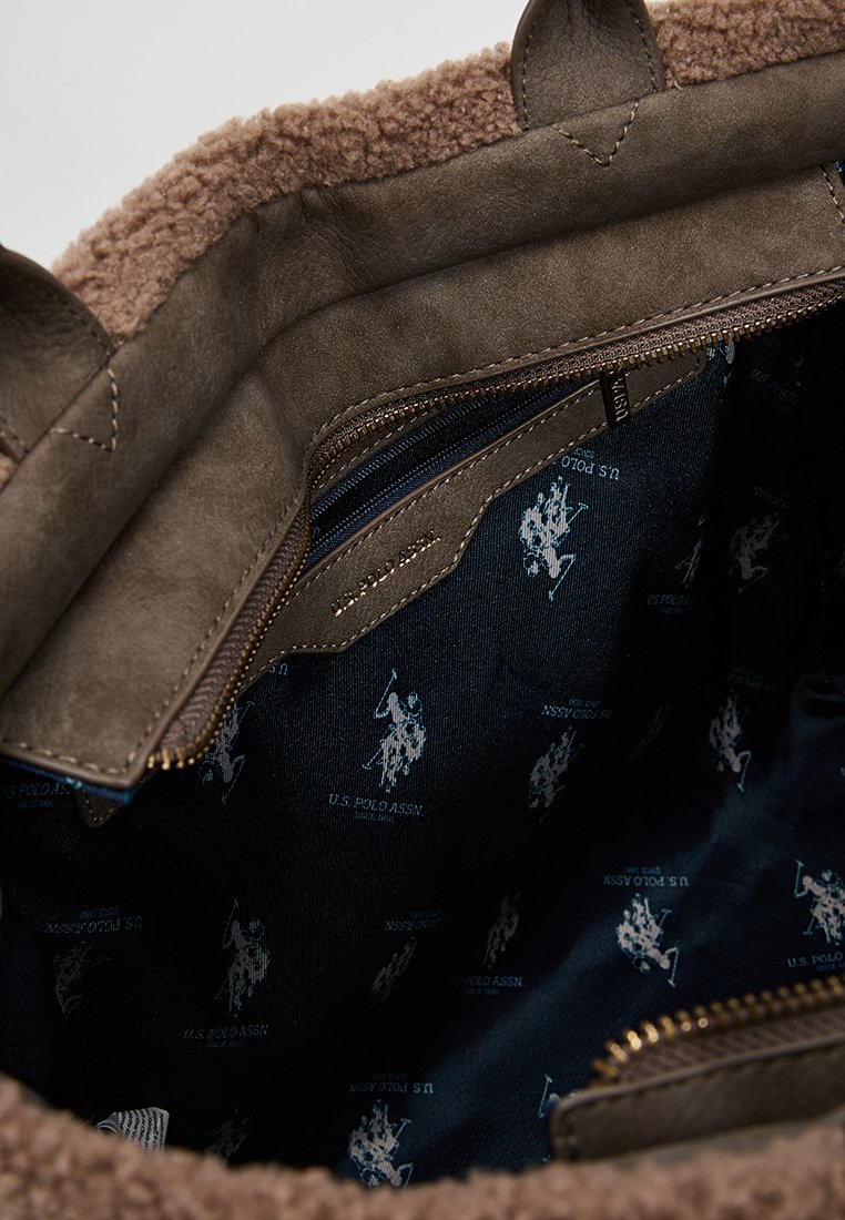 Intérieur d'un sac en faux shearling marron avec une doublure en tissu bleu marine, comportant de petits logos blancs U.S. Polo Assn. et une poche zippée. Accents en suède texturé.