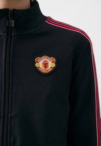 Črna jakna na zadrgo z visokim ovratnikom, ki ima rdeče in bele proge na ramenskih delih. Vezen logotip Manchester United na prsih.