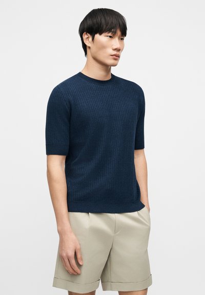 Pull tricoté bleu marine à manches courtes avec un motif côtelé texturé, encolure ronde et coupe décontractée, assorti à un short beige clair plissé.