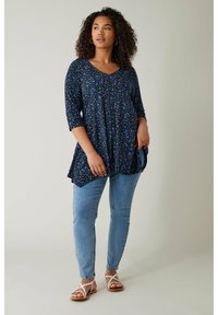 Schwarze Blumentunika mit blau-grünen Mustern, V-Ausschnitt, Dreiviertelärmel, kombiniert mit hellblauen Skinny Jeans und weißen Sandalen.