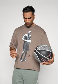 Pruun ülekaaluline t-särk must-valge graafikaga korvpallurist; tekstureeritud kangas, lühikesed varrukad, seljas rohelised lühikesed püksid ja käes korvpall.