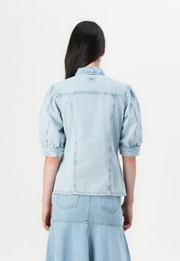 A woman with long dark hair wears a light blue denim jacket with puffed sleeves, paired with a matching denim skirt, facing away from the camera.