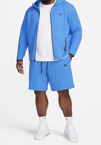 Man sombär en blå Nike zip-jacka och matchande shorts, vit Nike t-shirt, vita strumpor och vita sneakers, står med händerna i fickorna.