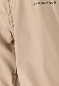 Veste d'extérieur beige avec une texture lisse, arborant un logo brodé en haut à gauche. Le tissu semble léger et flexible.