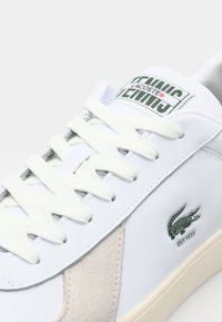 White leather sneaker with beige suede accents, textured laces, green logo, and vintage label on the tongue, designed for casual wear.