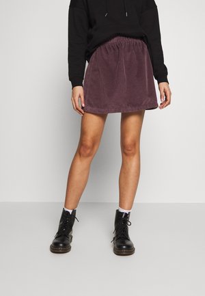 Femme portant une mini-jupe en velours côtelé violette, un sweat à capuche noir, des chaussettes blanches et des bottes noires à lacets, debout devant un fond gris.