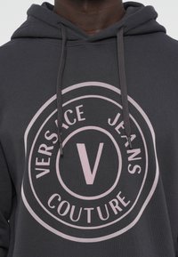 Charcoal sivá mikina s veľkým ružovým kruhovým logom, ktoré obsahuje nápis "VERSACE JEANS COUTURE" a centrálny dizajn písmena "V". Sťahovacie šnúrky ako dekorácia.