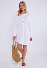 Robe chemise blanche à manches longues, en tissu texturé, avec cinq boutons. Le modèle tient un grand sac tissé couleur tan et porte des claquettes marron.