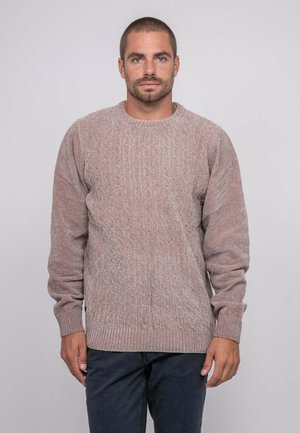 Homme aux cheveux courts et barbe portant un pull texturé en maille torsadée marron clair et un pantalon foncé, sur un fond uni clair.