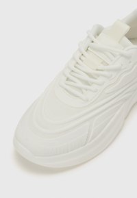 Weißer Sneaker mit einem strukturierten Mesh-Obermaterial, geschwungenen Designlinien und einer Gummisohle mit ausgeprägten Rillen. Die Schnürsenkel sind zentral und flach.