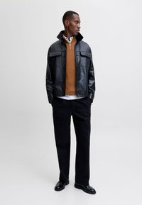 Giacca in pelle nera con chiusura a bottoni e due tasche sul petto, maglione marrone sotto, camicia a righe bianche, pantaloni neri e scarpe nere.