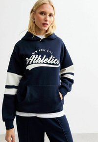 Marineblauwe hoodie met witte tekst "Athletics", contrasterende witte strepen op de mouwen, voorste kangoeroezak, ontspannen pasvorm en stoftextuur.