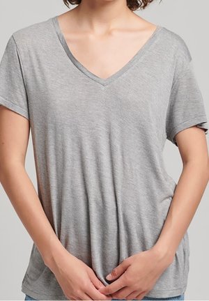 Woman wearing a loose-fitting, short-sleeve, light gray V-neck T-shirt, hands gently clasped in front at waist level.
