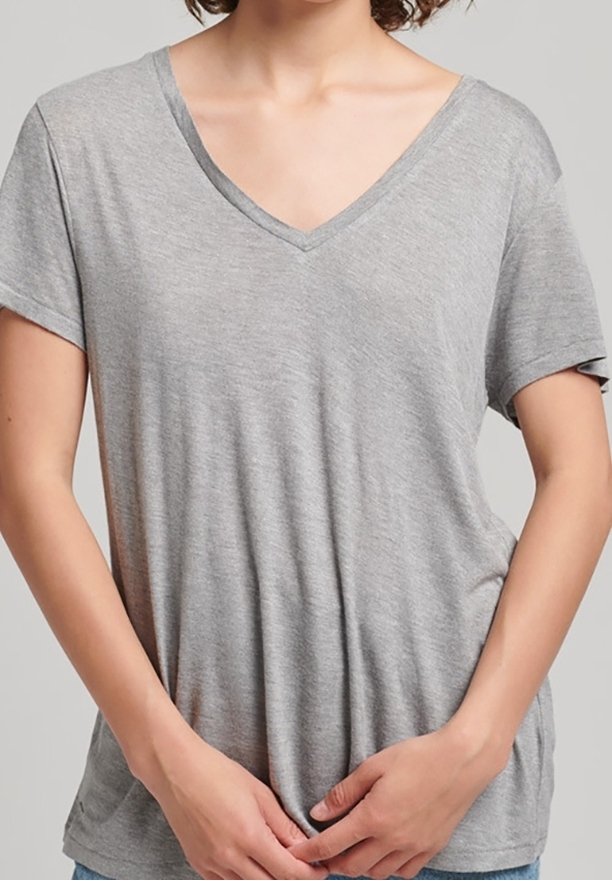 Femme portant un T-shirt ample à manches courtes et col en V de couleur gris clair, les mains doucement jointes devant à hauteur de taille.