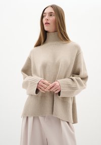 Beige pletený svetr s vysokým vroubkovaným límcem, �širokými rukávy a vroubkovaným lemem. Tkanina vypadá měkce a má texturu. V kombinaci se světlými kalhotami.