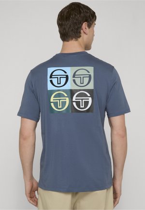 Mand iført en blå T-shirt med en grafik på ryggen bestående af fire firkanter, hver med en cirkel og en stiliseret "T"-logo i forskellige farver.