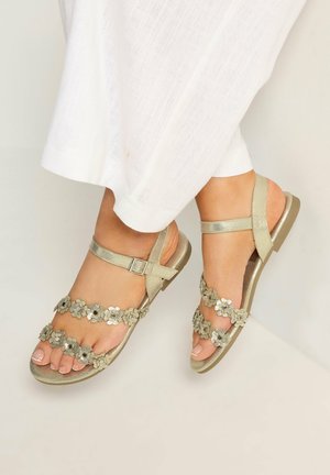 Füße in goldenen Sandalen mit Blumenmuster und Knöchelriemen, kombiniert mit weißen, strukturierten Hosen mit weitem Bein.