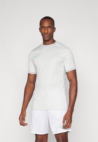 Camiseta atlética Nike de color gris claro con mangas cortas, cuello redondo y logo de acento en color azul verdoso. Combinada con pantalones cortos blancos con estampado. Textura de tela suave.