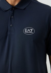 Tums zilo polo krekls, kas izgatavots no teksturēta auduma, ar trīs pogām un baltu izšuvumu ar EA7 Emporio Armani logo uz krūšu.