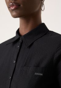 Svart teksturert skjorte med krage, frontlomme og en liten Calvin Klein-lapp på lommen.