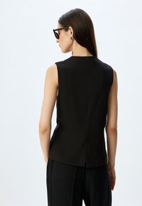 Schwarzes ärmelloses Oberteil mit hohem Kragen und Rückenbelüftung, aus glattem Material gefertigt. Das Modell trägt schwarze Hosen und dunkle Sonnenbrillen.