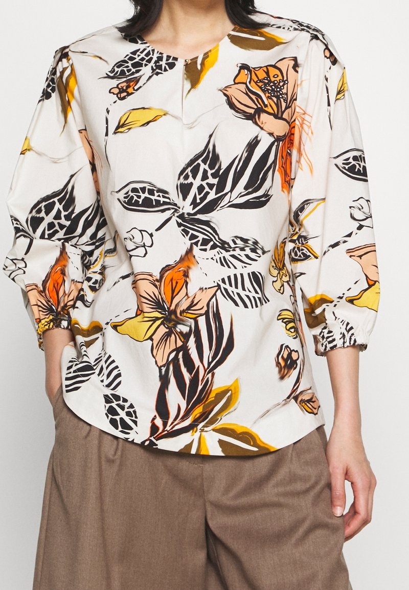 Blouse blanche à imprimé floral orange, jaune et noir, manches trois-quarts bouffantes, portée avec un pantalon marron ample.