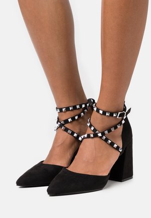 Chaussures à talons hauts en suede noir avec une pointe aiguille, dotées de sangles croisées ornées de clous en forme de pyramide argentée et de talons carrés.