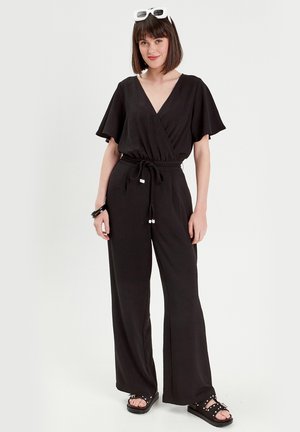 Mono negro con escote en V, mangas cortas de vuelvo, cintura elástica y patas anchas. Combinado con sandalias negras y gafas de sol blancas de gran tamaño.