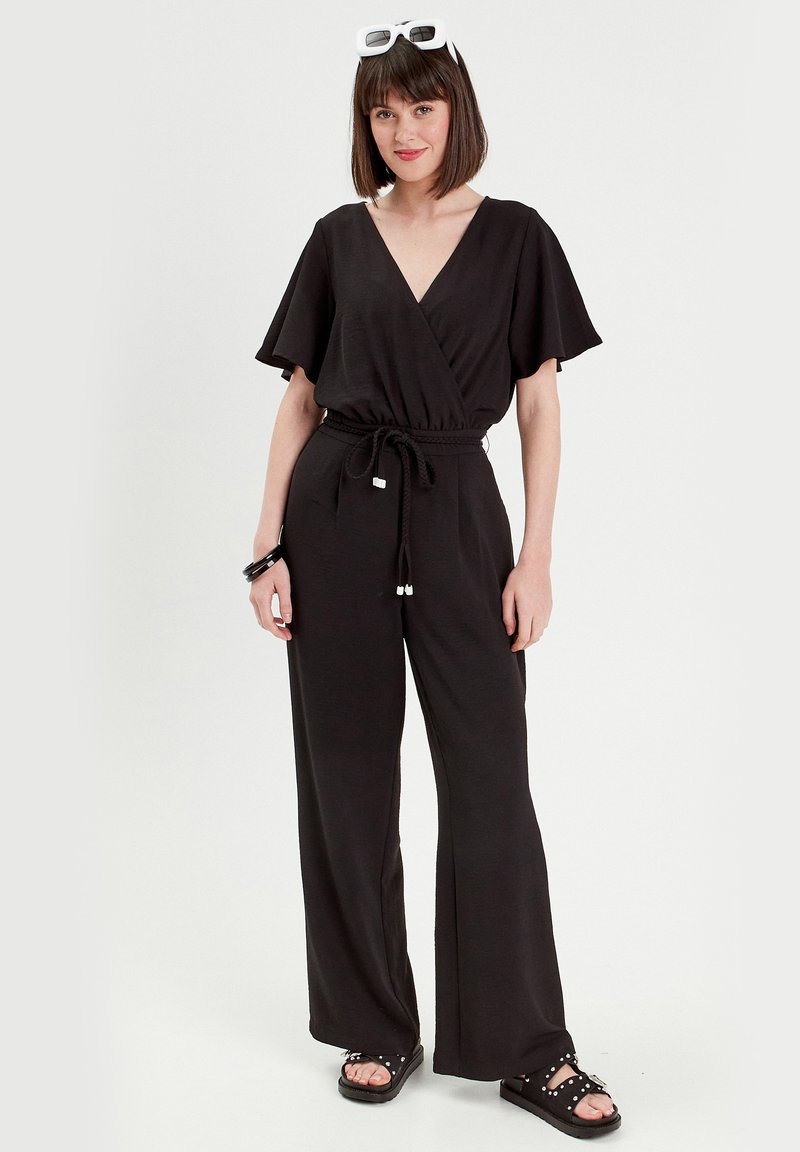Schwarzer Jumpsuit mit V-Ausschnitt, kurzen, schwingenden Ärmeln, elastischem Taillenbund und weiten Beinen. Kombiniert mit schwarzen Sandalen und übergroßen weißen Sonnenbrillen.
