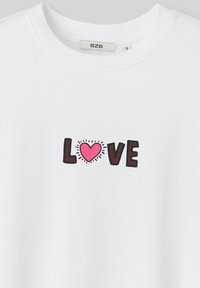 T-shirt en coton blanc avec le mot "AMOUR" en lettres noires épaisses, accompagné d'un cœur rose et de lignes rayonnantes autour.