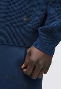 Kötött kék pulóver texturált mintával és bordázott mandzsettákkal, sötétkék nadrággal párosítva, amelyen oldalán finom „BOSS” logó látható.