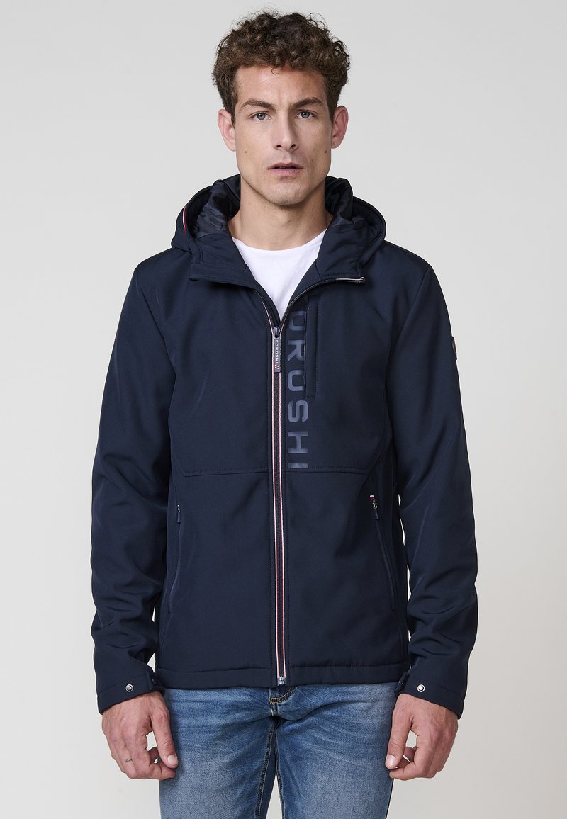 Chaqueta azul marino con cremallera frontal, capucha ajustable y detalle de logo, con una textura suave y dos bolsillos laterales.