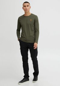 Olive green long-sleeve shirt, crew neckline, with a small orange logo. Worn with black cargo pants and black sneakers.