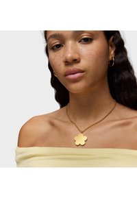 Ciondolo a forma di fiore in oro su una catena fine, indossato su spalle scoperte, abbinato a un top giallo chiaro a spalle scoperte. Superficie liscia e lucida.