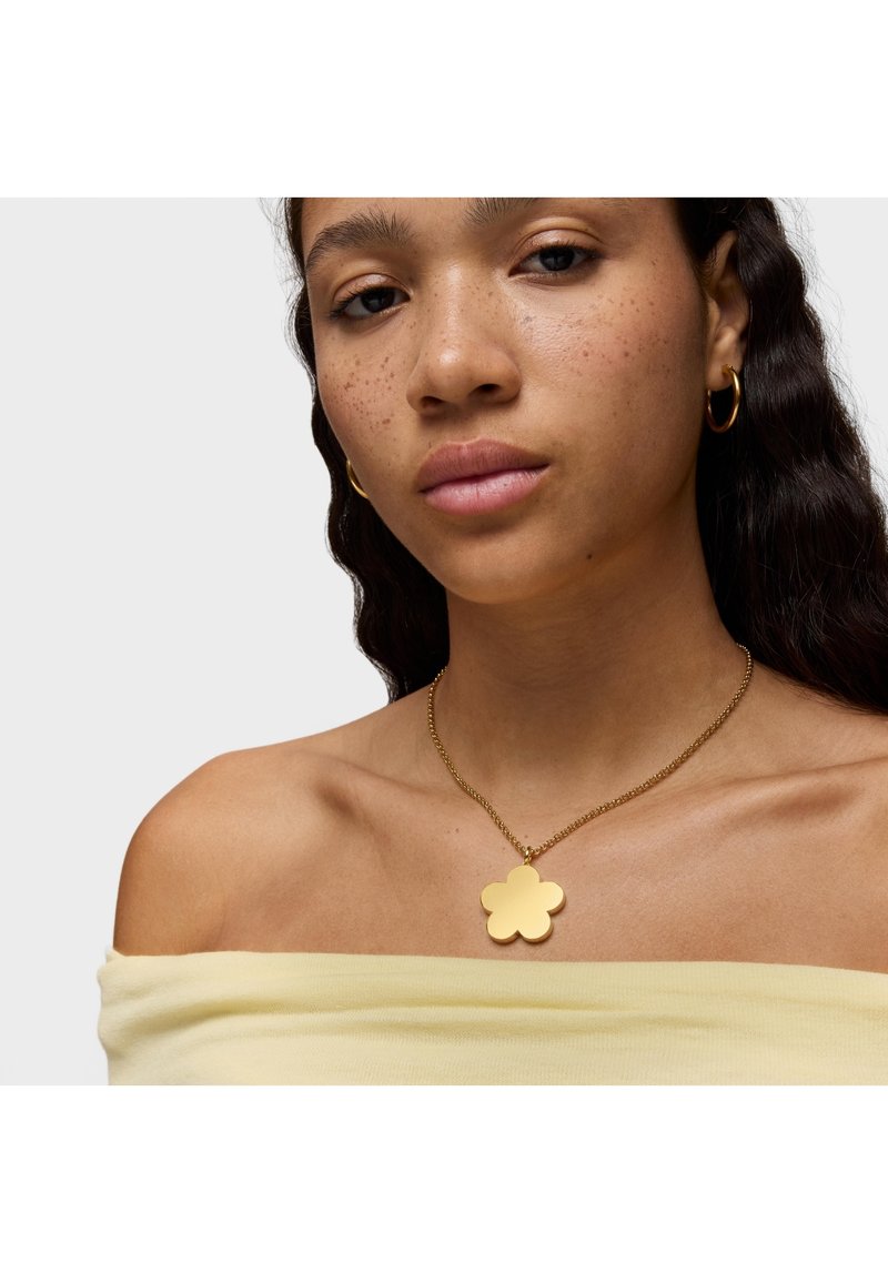 Ciondolo a forma di fiore in oro su una catena fine, indossato su spalle scoperte, abbinato a un top giallo chiaro a spalle scoperte. Superficie liscia e lucida.