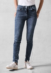 Jeans ajustados de denim azul oscuro, con un diseño de cinco bolsillos, cierre de botón y una textura suave. Combinados con zapatillas blancas.