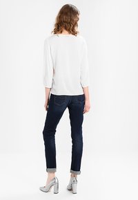 Blusa blanca con mangas de tres cuartos, combinada con jeans azul oscuro arremangados en los puños y zapatos de tacón plateados, vista desde atrás.