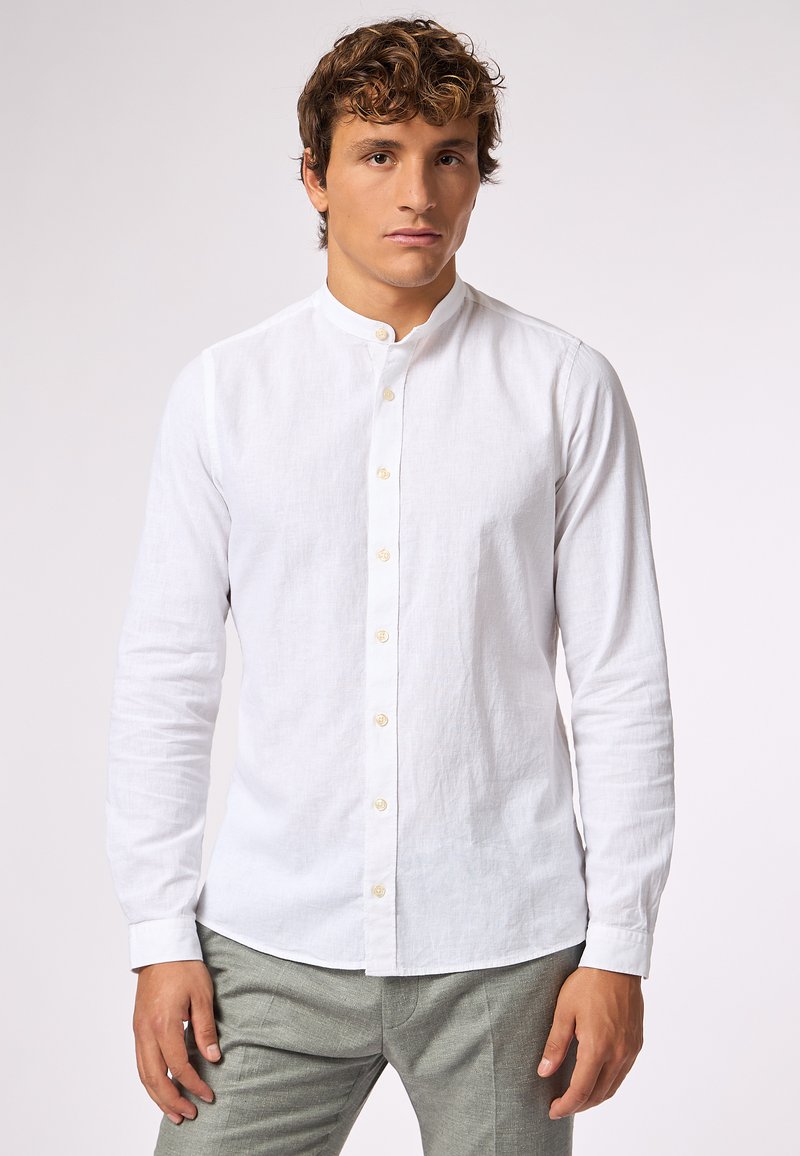 Camicia bianca in lino con colletto alla coreana, maniche lunghe e bottoni beige, abbinata a pantaloni grigi chiari. Texture liscia e design classico.