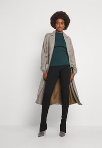 Um casaco xadrez longo em bege e preto, usado sobre uma blusa canelada em azul escuro, combinado com calças pretas justas e sapatos de salto alto cinzas.