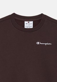 T-shirt in cotone marrone con collo rotondo e maniche corte. Presenta un logo Champion ricamato in bianco sul petto. Texture liscia.