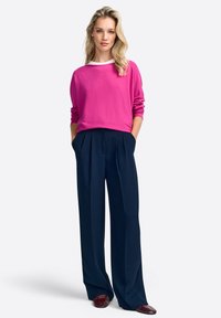 Roze gebreide trui met een witte kraag, gecombineerd met marineblauwe broek met wijde pijpen, voorzien van plooien en zijzakken, aangevuld met bordeauxrode schoenen.