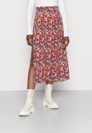 Femme portant une jupe midi rouge à fleurs avec une fente latérale, un haut blanc à manches longues et des bottines blanches épaisses, debout sur un sol gris.