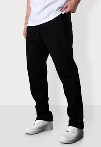 Schwarze Sweatpants mit einem Kordelzugbund, Seitentaschen und Druckknopfverschlüssen an den Knöcheln, kombiniert mit weißen Sneakers. Weicher, glatter Stoff.