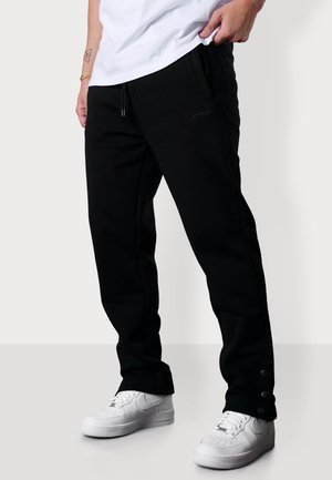 Black sweatpants with a drawstring waist, side pockets, and snap closures at the ankles, paired with white sneakers. Soft, smooth fabric.