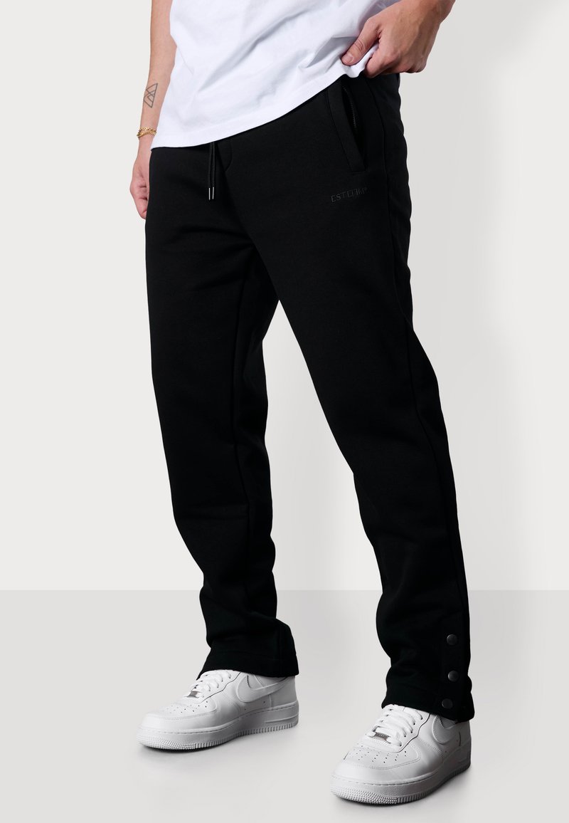 Schwarze Sweatpants mit einem Kordelzugbund, Seitentaschen und Druckknopfverschlüssen an den Knöcheln, kombiniert mit weißen Sneakers. Weicher, glatter Stoff.