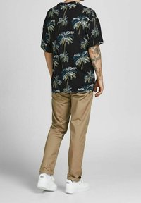 Korte mouwen zwart shirt met groene en blauwe palmboomprint, gecombineerd met beige broeken en witte sneakers. Zichtbare tatoeage op de arm.